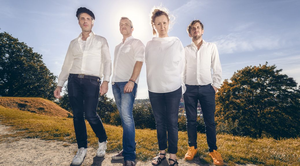 Thymeshift + Amsler. Porträtt i motljus med gruppen. Med solen i motljus bakom sig står gruppens fyra medlemmar med vita skjortor och jeans i en sommrig miljö.