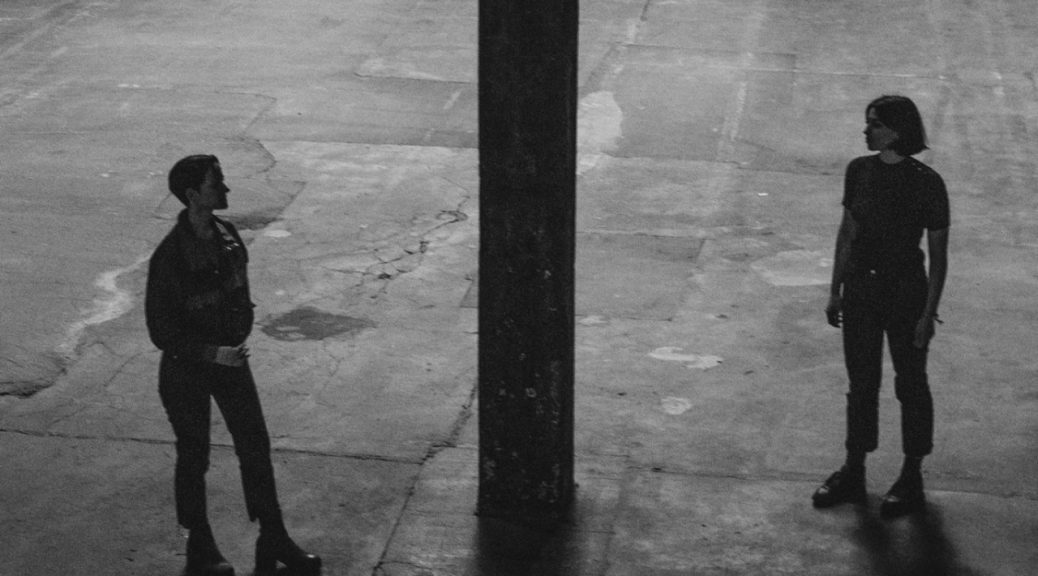 upper glossa. de två bandmedlemmarna står på var sin sida av en pelare i en industrilokal. de tittar på varandra. svartvit bild, en rå känsla.